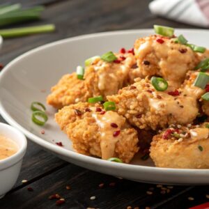 Golden, crispy Bang Bang Chicken bites drizzled with creamy, spicy sauce, garnished with green onions and sesame seeds, served on a white plate.